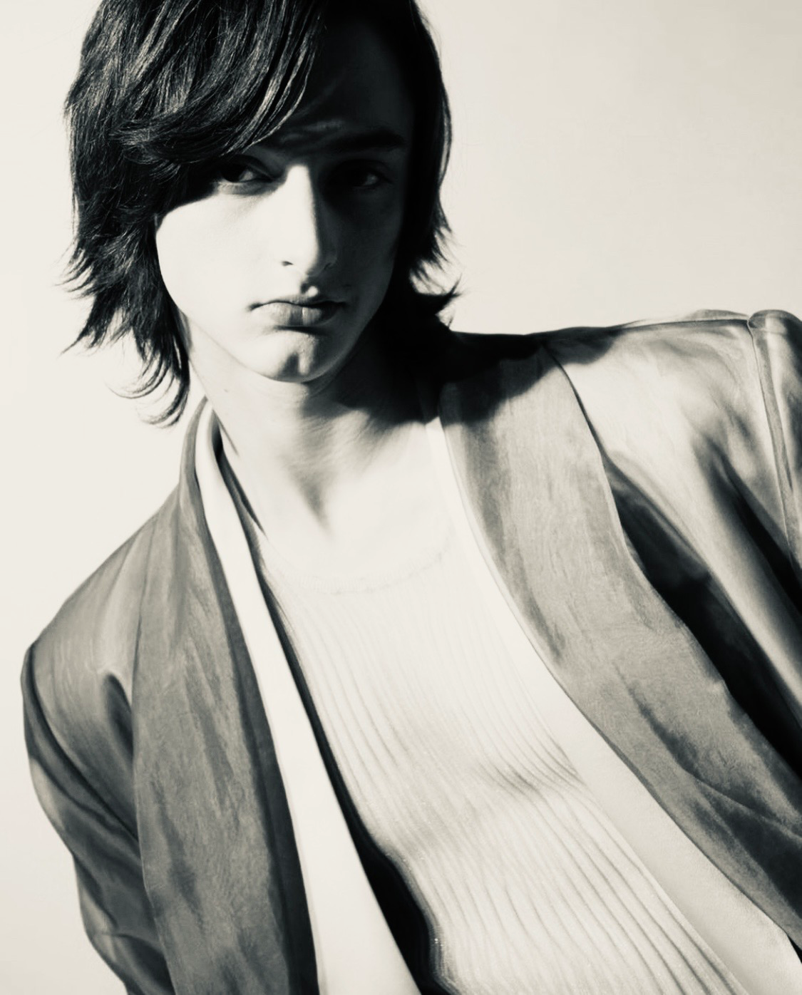 Black-and-white portrait of a young person with chin-length dark hair, wearing a light jacket over a ribbed shirt. The subject looks slightly to the side with a calm, composed expression.