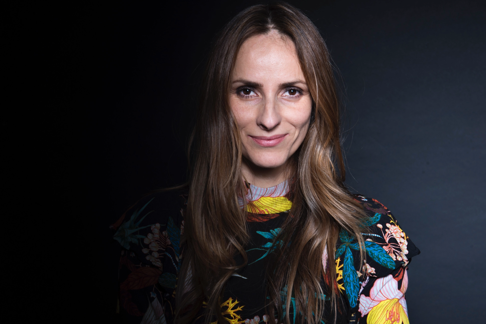 professional headshot of a woman with long straight brown hair, wearing a vibrant floral-patterned top, smiling softly against a dark studio background. Movement director.