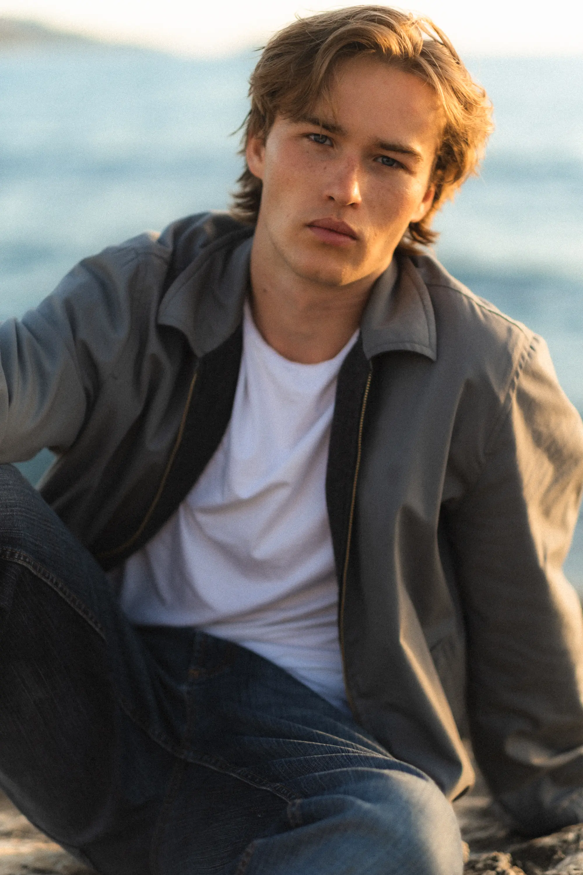 Young man in a grey jacket and white tee seated casually on the beach – effortless editorial style with natural light and an ocean backdrop.