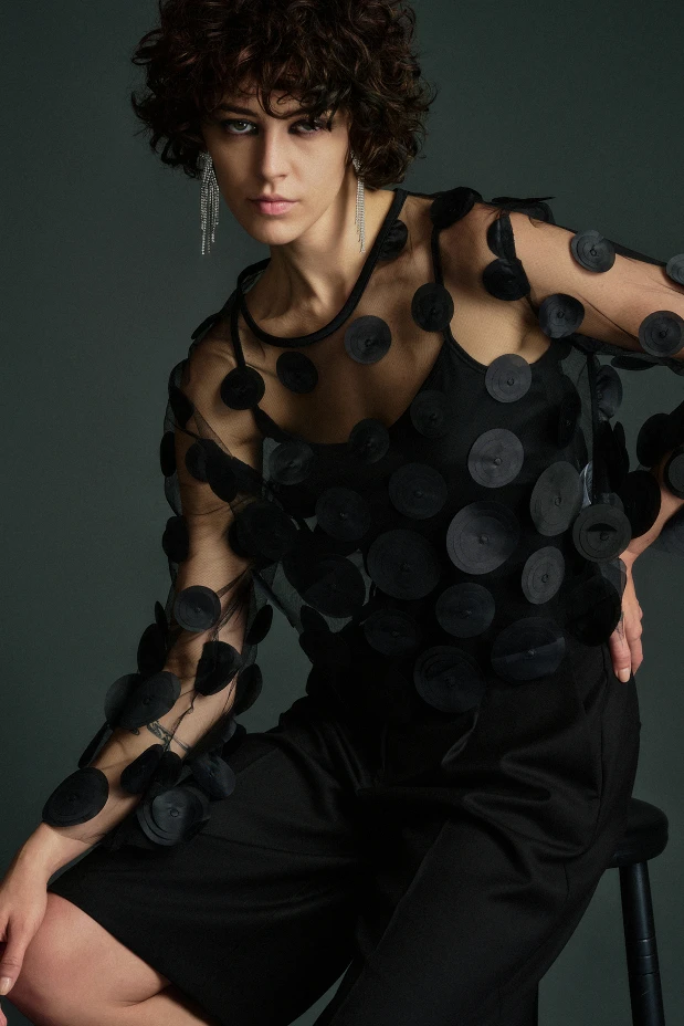 Woman wearing a sheer black top with bold 3D circular appliqués over a black outfit, styled with dangling crystal earrings – dramatic editorial fashion against a dark green background.