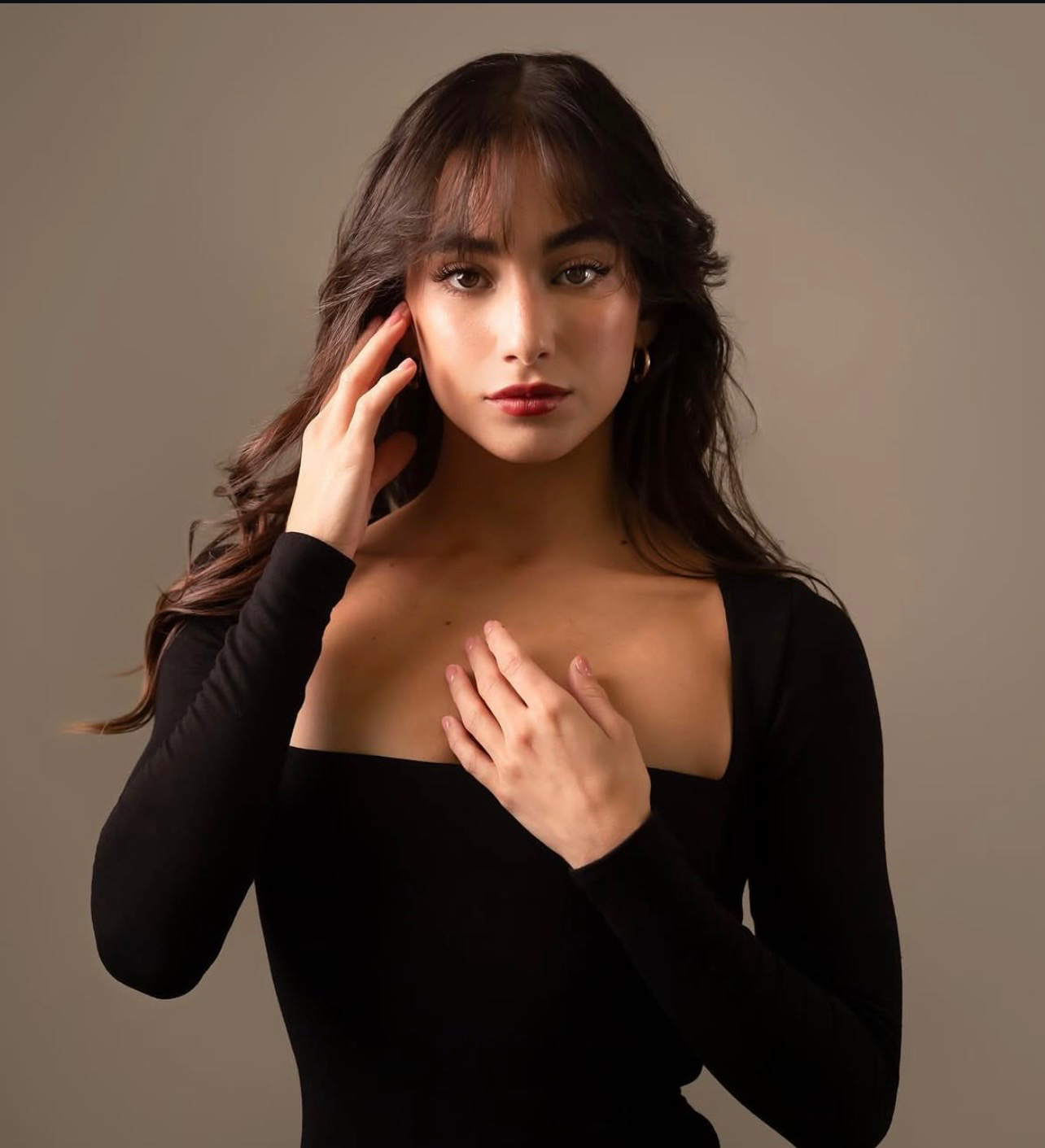Studio portrait of a dark-haired woman in a black off-shoulder top, hand raised to her cheek, against a neutral warm-grey backdrop.