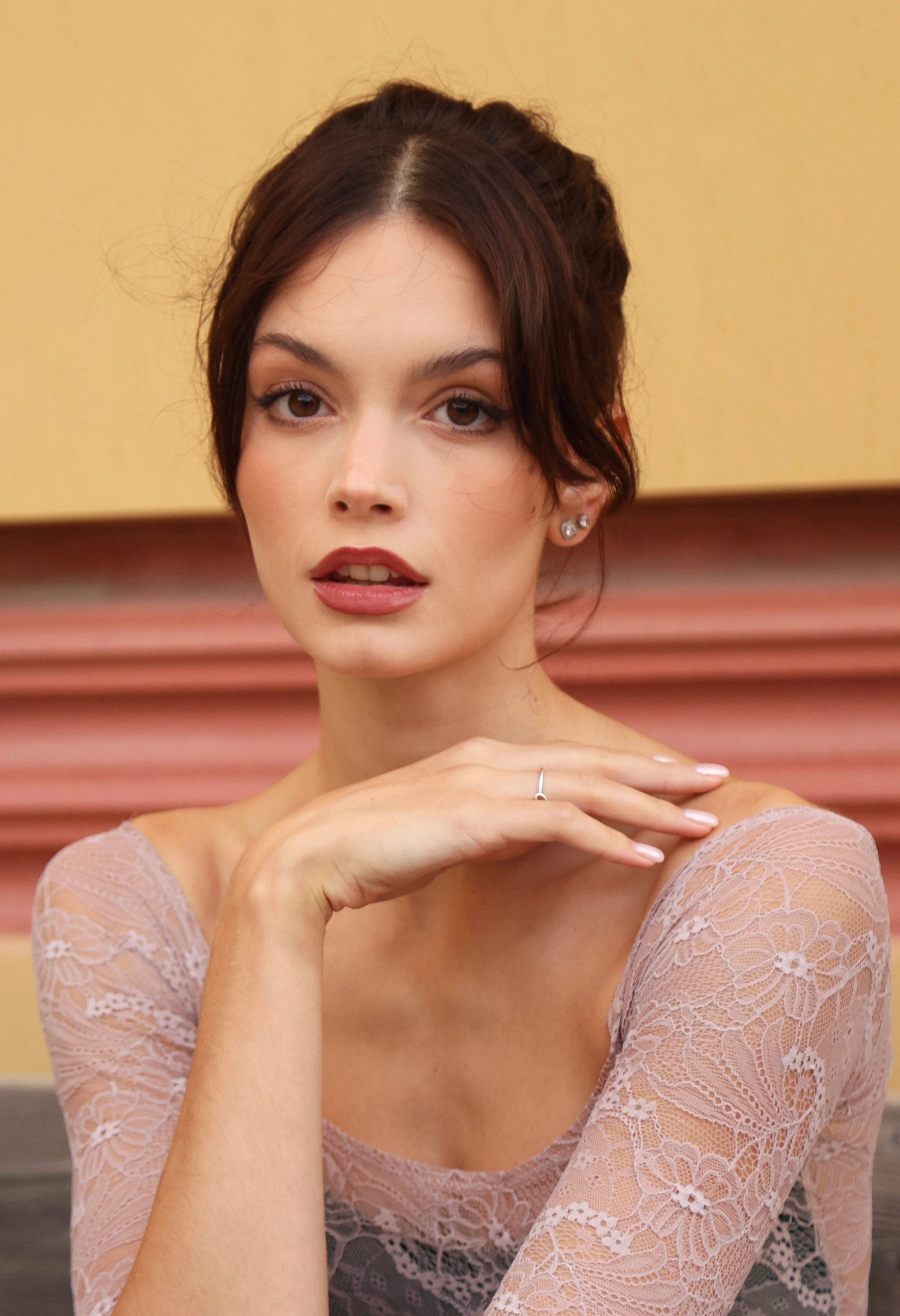 Portrait of a brunette woman with her hair in an updo, wearing a dusty pink lace top, resting her chin on her hand, against a warm yellow and terracotta backdrop.