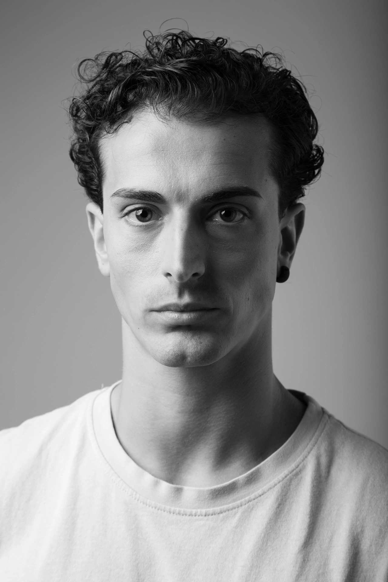 Schwarz-weißes Studioporträt eines jungen Mannes mit dunklen Locken in einem weißen T-Shirt, direkt in die Kamera blickend, vor neutralem grauem Hintergrund.