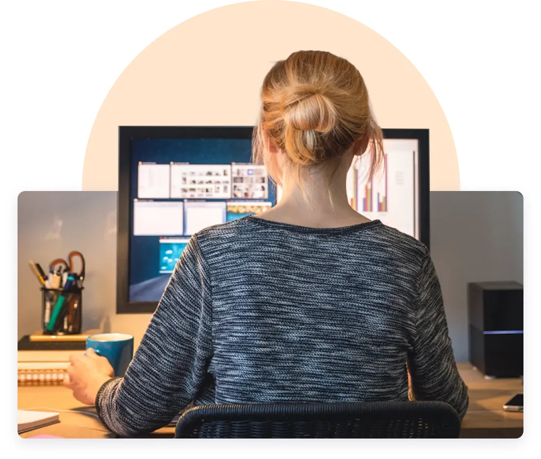 A woman with a bun hairstyle sitting at a desk, working on a computer with multiple windows open on the screen. She holds a blue mug, with office supplies to her left and a light beige background enveloping the top part of the image.