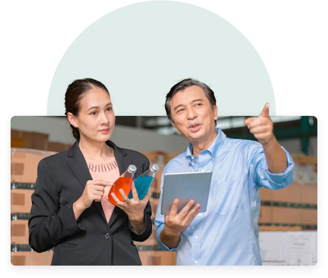 A woman in a black blazer holding two bottles of colored liquid listens attentively to a man in a light blue shirt pointing forward, with a tablet in his other hand, set against a pale blue semi-circular background with warehouse boxes in the backdrop.