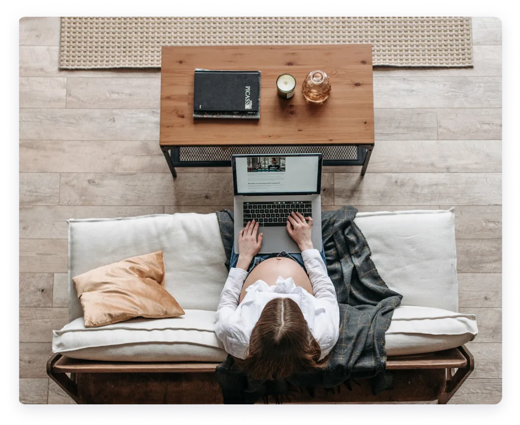The image shows a top-down view of a pregnant person working on a laptop. The person is seated on a comfortable couch or daybed with light-colored cushions.