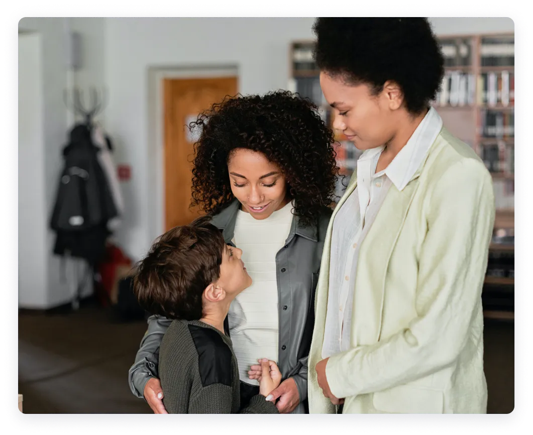 Two women smiling and looking at a young boy in an indoor setting.