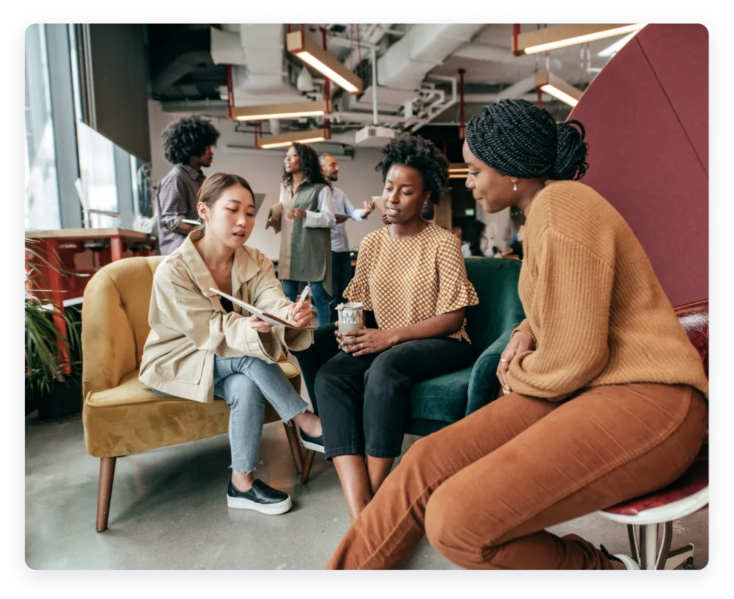 Three women engaged in a conversation in a modern, open space. Other individuals can be seen in the background, giving a feel of a collaborative environment.