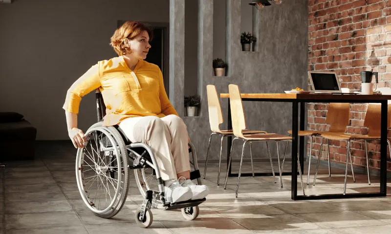 Redhead woman in a wheelchair wearing a yellow shirt and tan pants. She is looking at a brick wall.