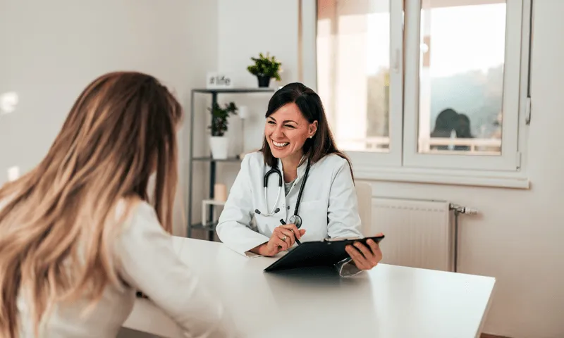 White female doctor with brown hair smiling and speaking to a patient. The patient's face is not shown.