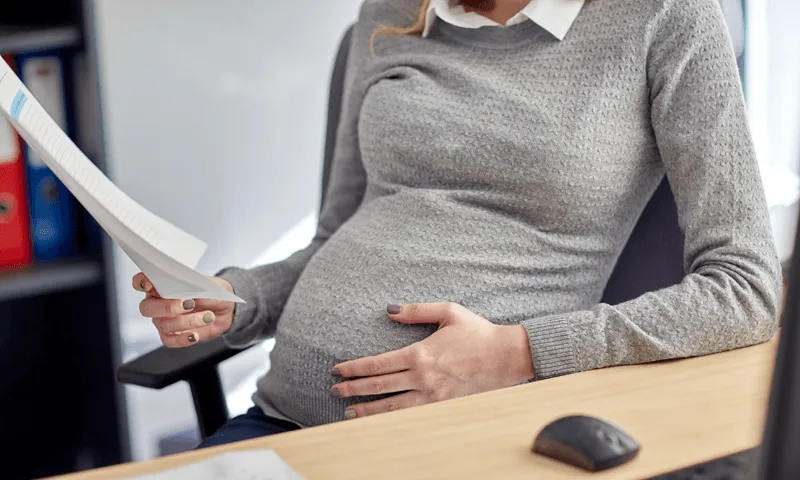 Pregnant woman sitting at desk holding baby bump. Wearing a grey sweater. 