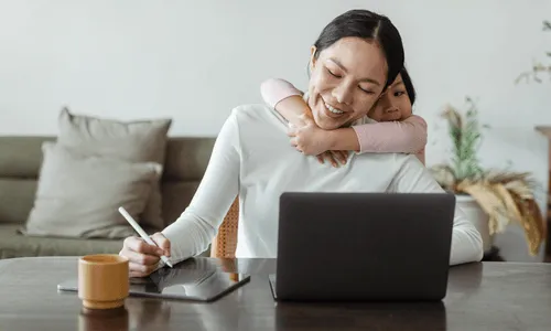 Woman working on her computer with a pen in her hand. Her daughter is hugging her behind her neck.