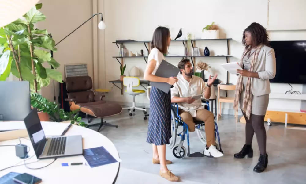Two women are standing next to a man in a wheelchair. They are conversing in the office.