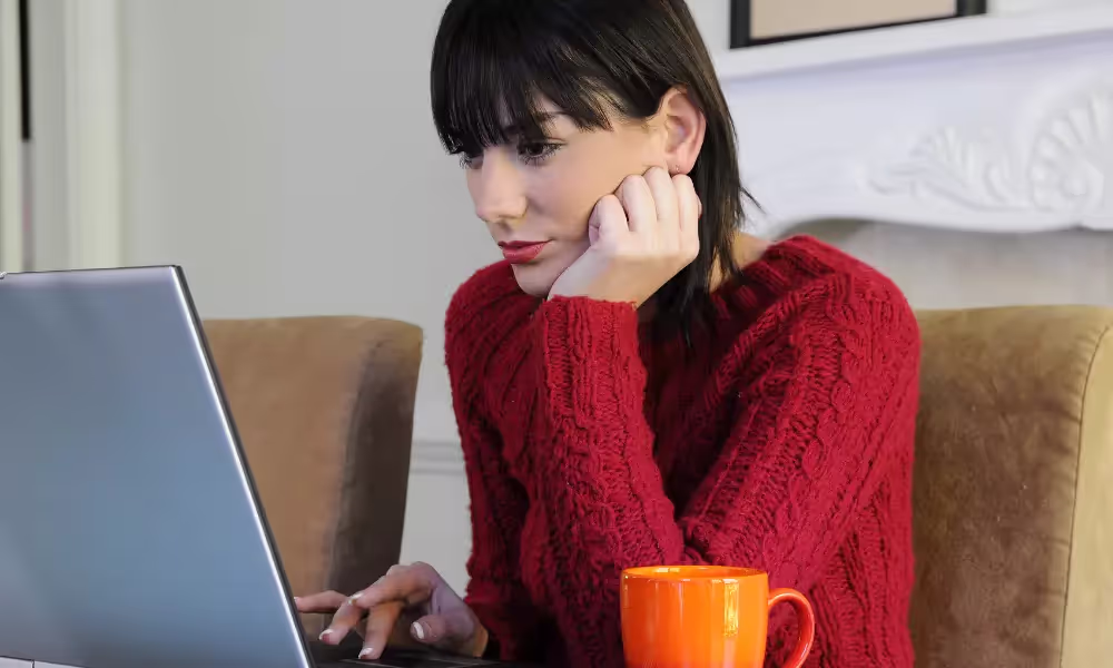 Woman with black hair and bangs wearing a red sweater. She is looking at her computer and has an orange coffee cup on her desk.
