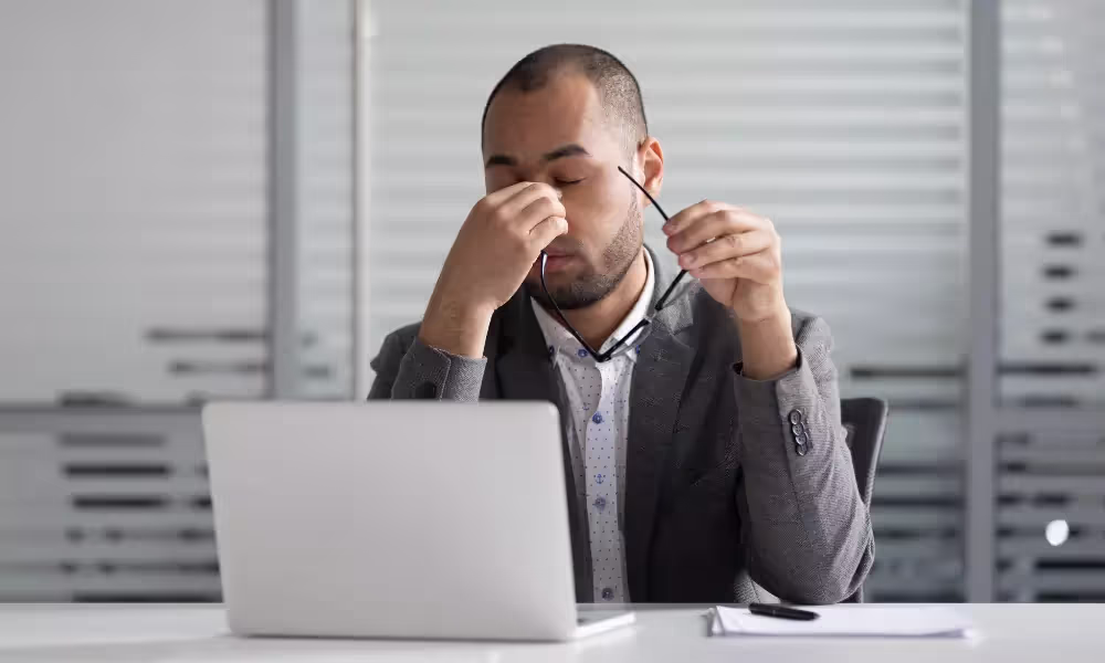 Man holding glasses and rubbing face behind computer screen
