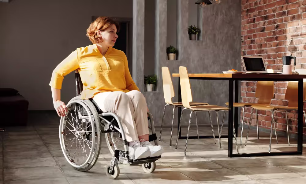 Redhead woman in a wheelchair wearing a yellow shirt and tan pants. She is looking at a brick wall. 