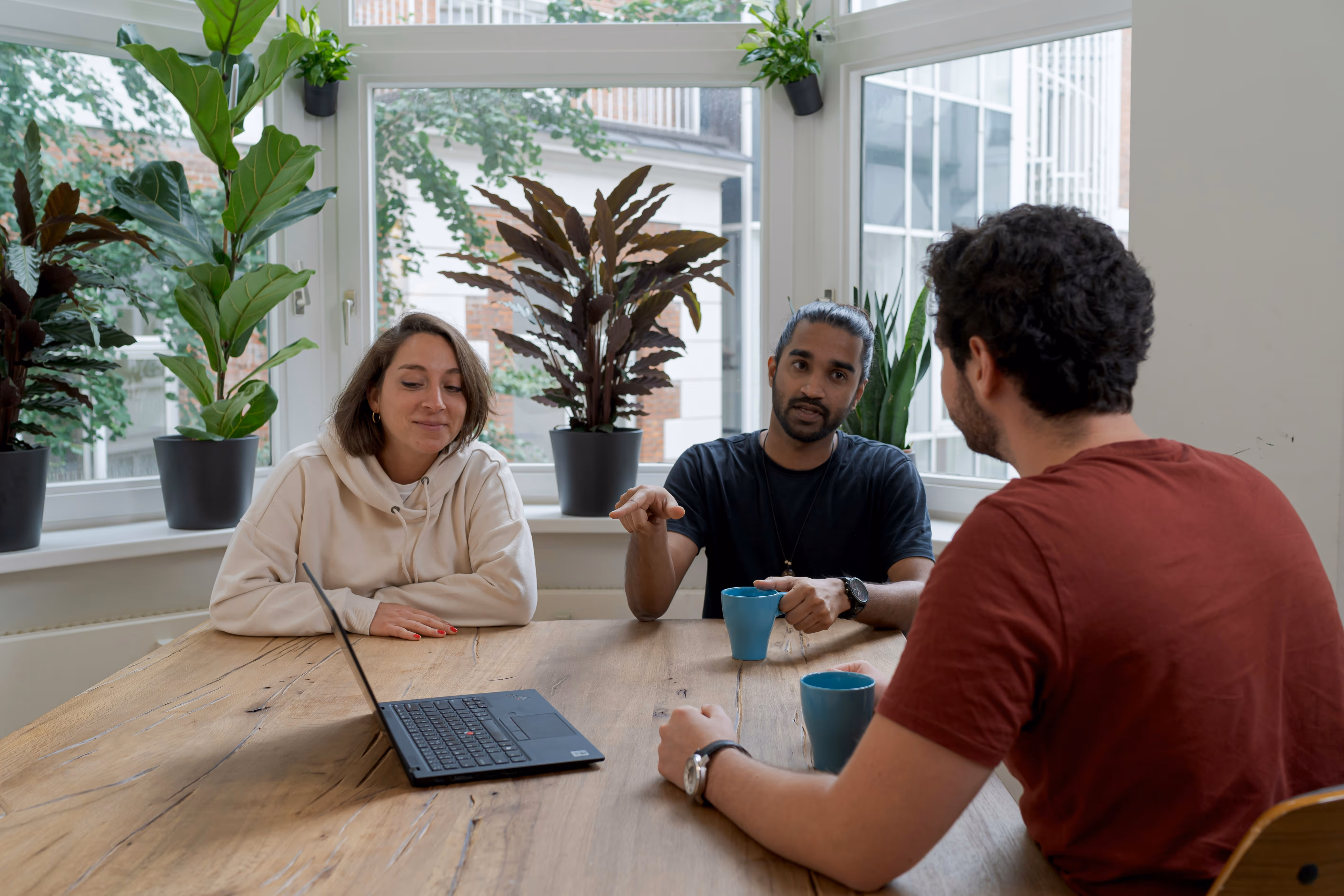 Three people having a discussion around a wooden table with a laptop and coffee mugs, sitting in front of a large window with green plants.