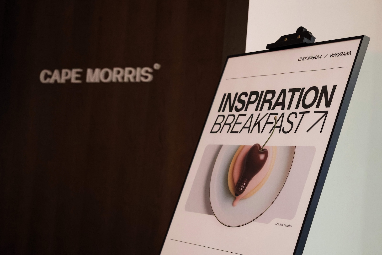 Zdjęcie przedstawia sztalugę z tablicą na które jest nazwa i logo wydarzenia. W tle widoczne jest logo firmy Cape Morris.