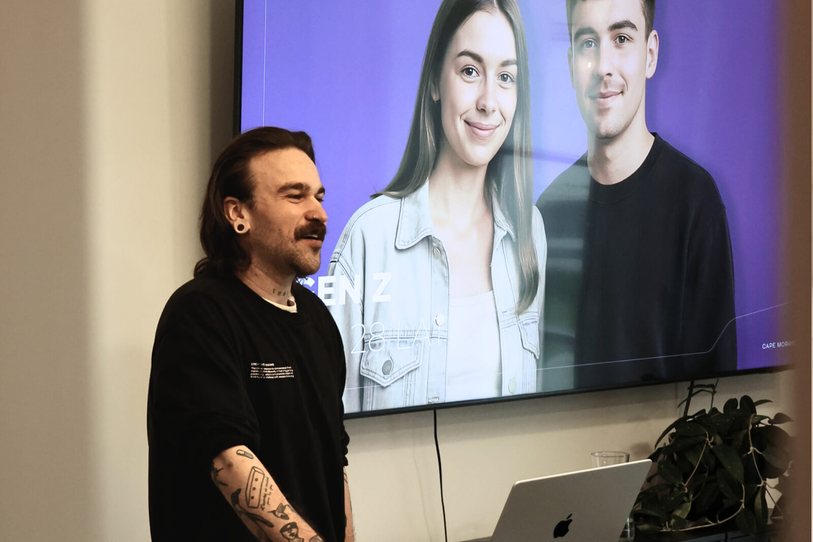 Zdjęcie przedstawia jednego z prowadzących Pawła w trakcie prezentacji. Na ekranie za nim jest zdjęcie z prezentacji przedstawiające dwoje młodych ludzi.