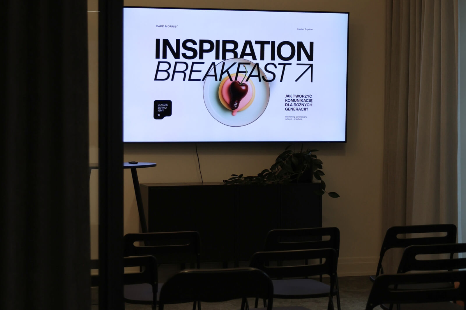 Zdjęcie przedstawia salę w której odbywa się wydarzenie. Przed ekranem na którym wyświetla się logo i nazwa wydarzenia s ustawione czarne składane krzesła.