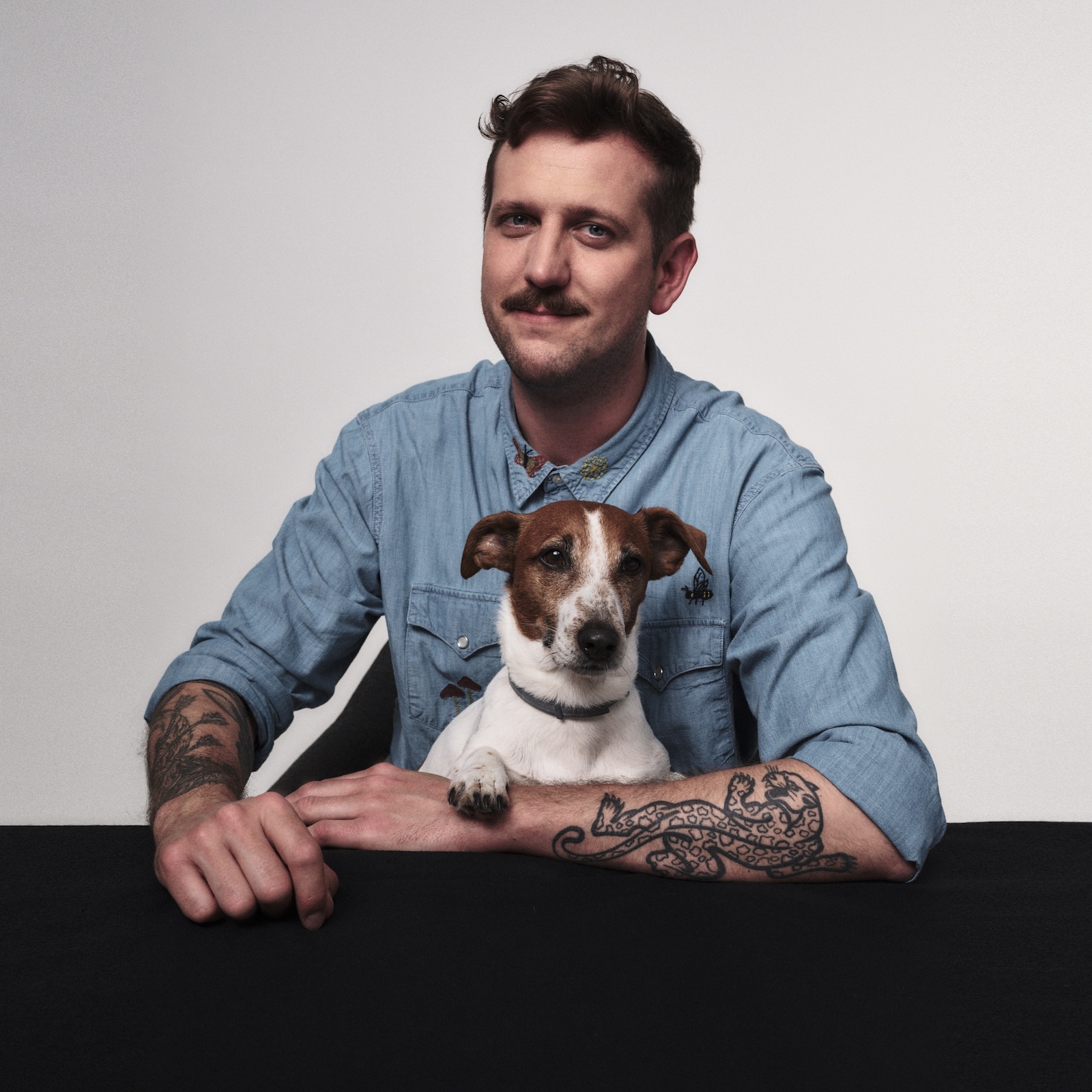 Zdjęcie przedstawia osobę odpowiedzialną za kontakt z uczestnikami oraz jednego z prowadzących Krzysztofa Żukowskiego. Krzysztof siedzi przy stole a na jego kolanach znajduje się nasz Senior Good Boy, piesek Stefan.