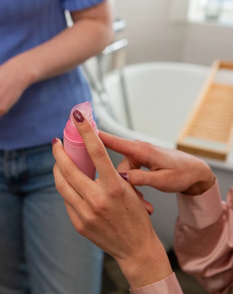 Fokus på en persons händer med välskötta lila naglar som håller i en liten rosa plastflaska med en pumpdispenser. En hand håller flaskan medan den andra tar av locket.