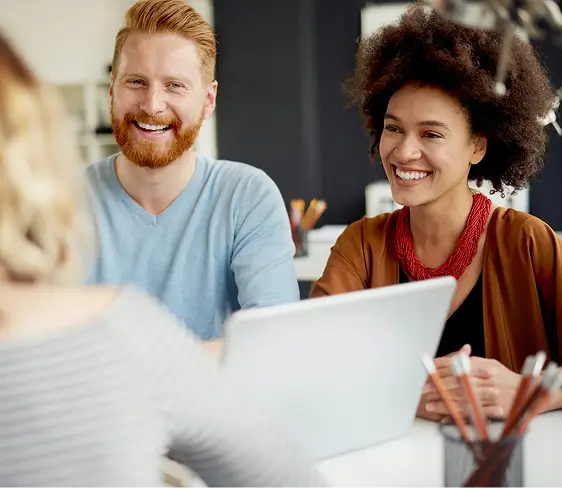 Two smiling colleagues, a red-haired man and a woman with curly hair and a red necklace, sitting at a desk with a laptop, engaged in a friendly conversation with a third person.