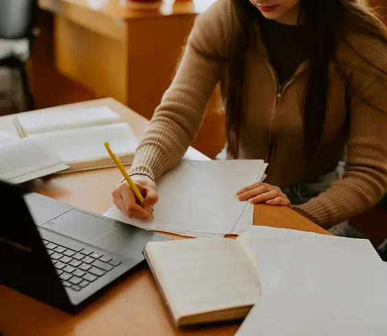 Person writing on papers at a desk with a laptop and open books nearby.