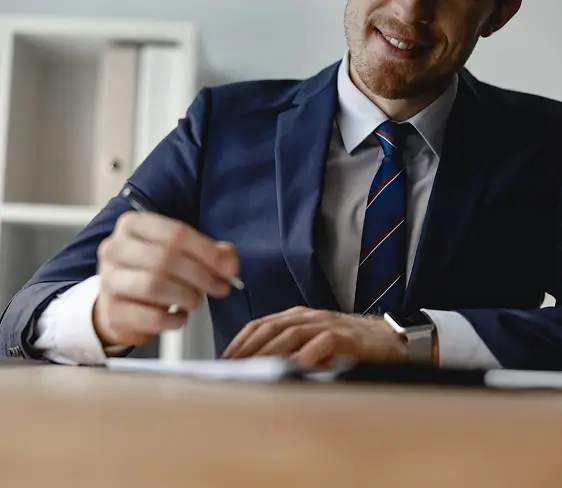 Smiling businessman in a blue suit and striped tie writing on paper at a desk.