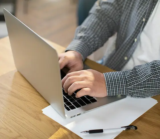 Person typing on a laptop placed on a wooden table with a pen and blank papers.