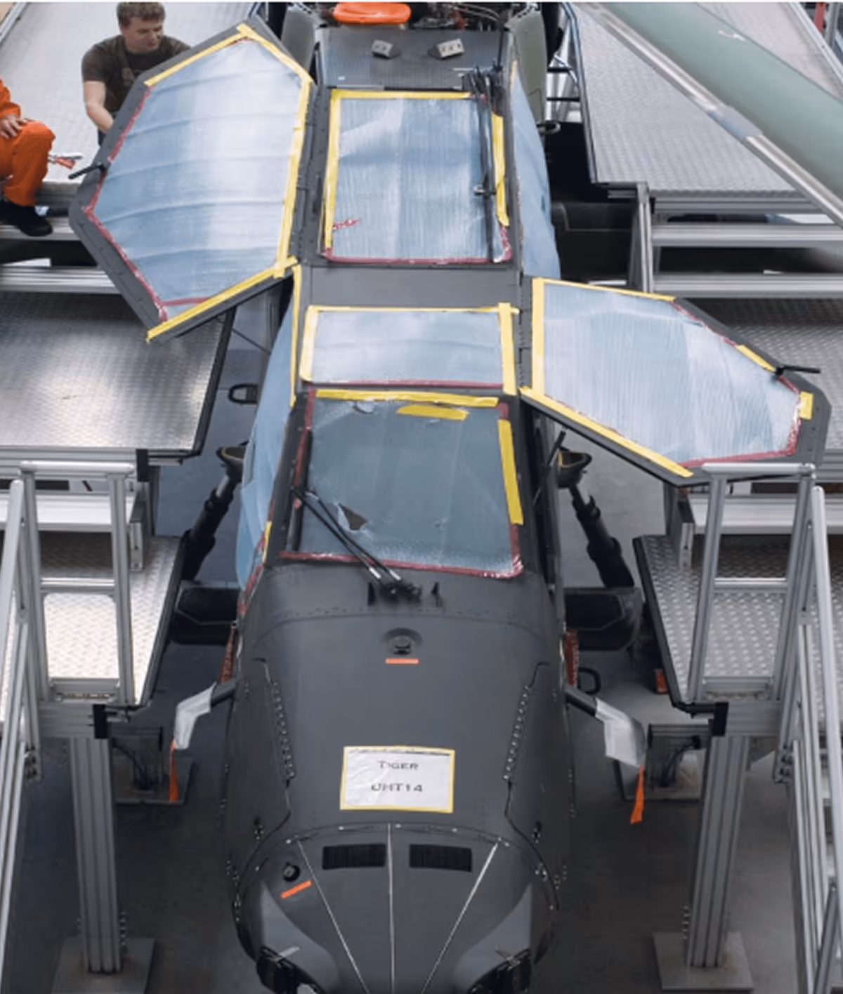 Helikopter im Hangar mit geöffneten Wartungsklappen auf Plattformen, Techniker im Hintergrund für Inspektion.