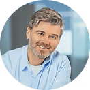 Freundlich lächelnder Mann mit grauen Haaren und Bart in einem hellblauen Hemd, vor unscharfem Bürohintergrund.