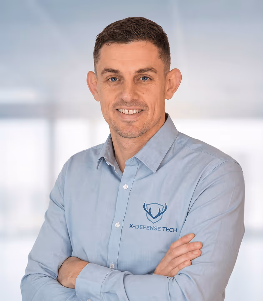 Lächelnder Mann in blauem Hemd mit K-Defense Tech Logo, steht mit verschränkten Armen vor einem unscharfen Bürohintergrund.