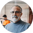 Älterer Mann mit Brille und Bart in grauem Pullover vor unscharfem Hintergrund in Büro.