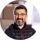 Mann mit Brille und Bart lächelt, trägt schwarze Daunenweste; unscharfer, heller Hintergrund.