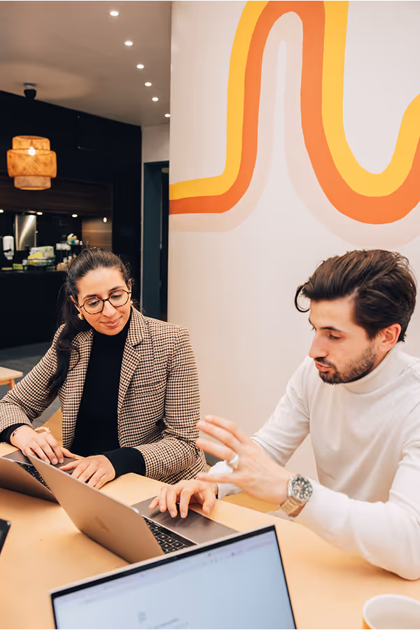 Deux collègues assis à une table discutant avec des ordinateurs portables ouverts devant eux dans un bureau moderne.