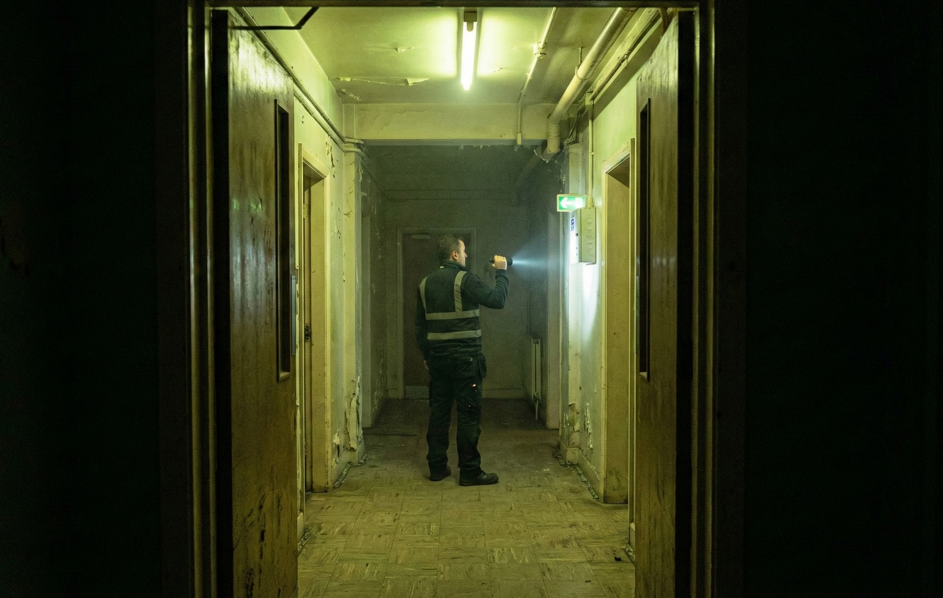 A man in dark clothing holding a flashlight shines light on a wall in a dim hallway with peeling paint and worn floors.