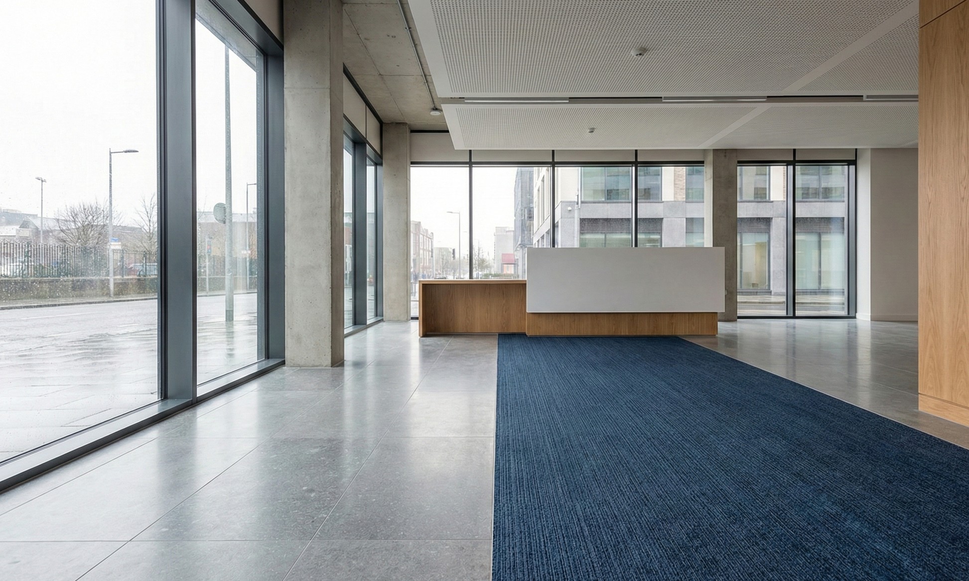Modern office lobby with large glass windows, concrete walls, gray tile floor, a wooden reception desk with white front panel, and a blue carpet runner.