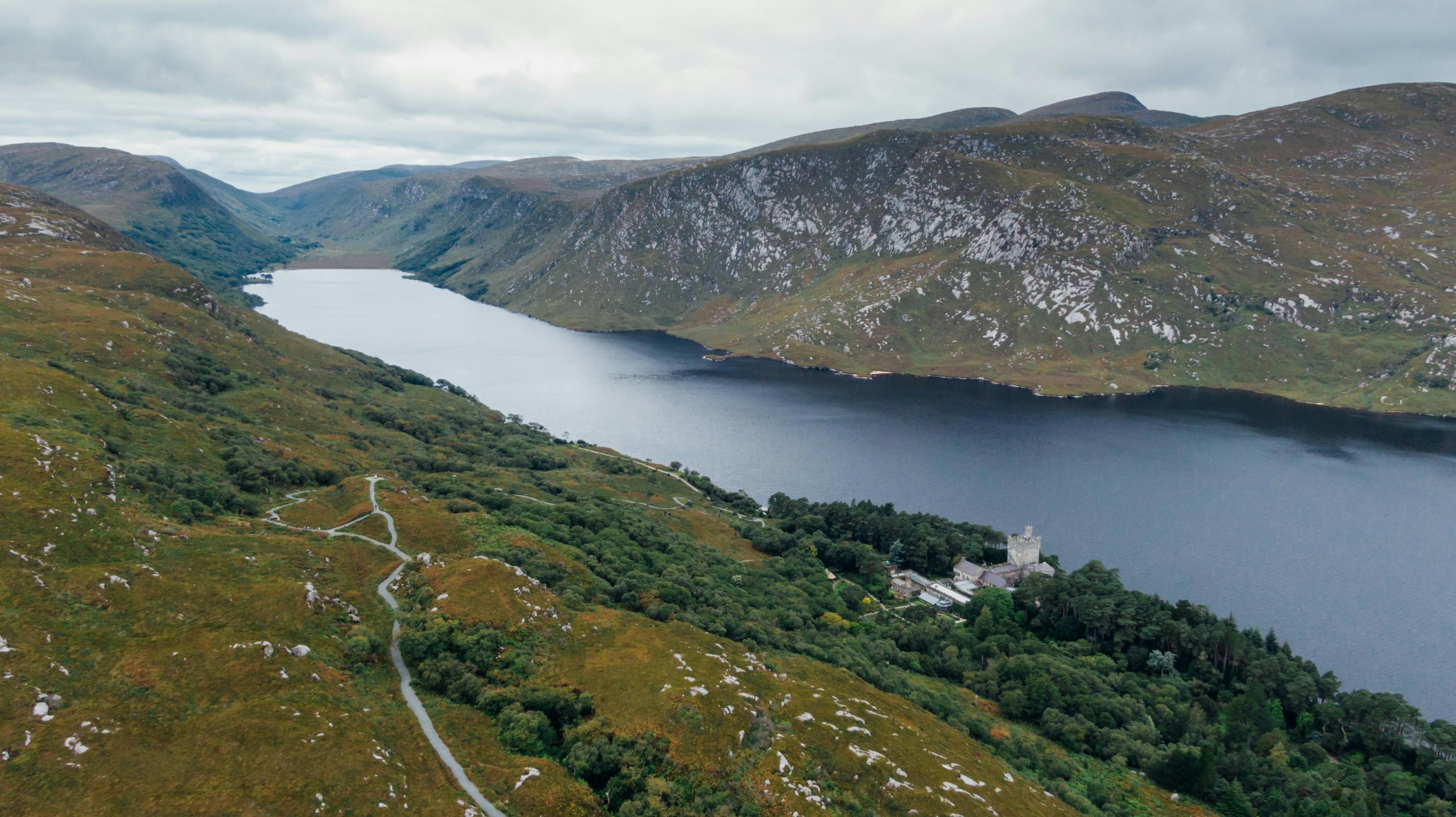 Aerial view of a narrow lake surrounded by green hills and rocky mountains under a cloudy sky, with a castle near the water's edge.