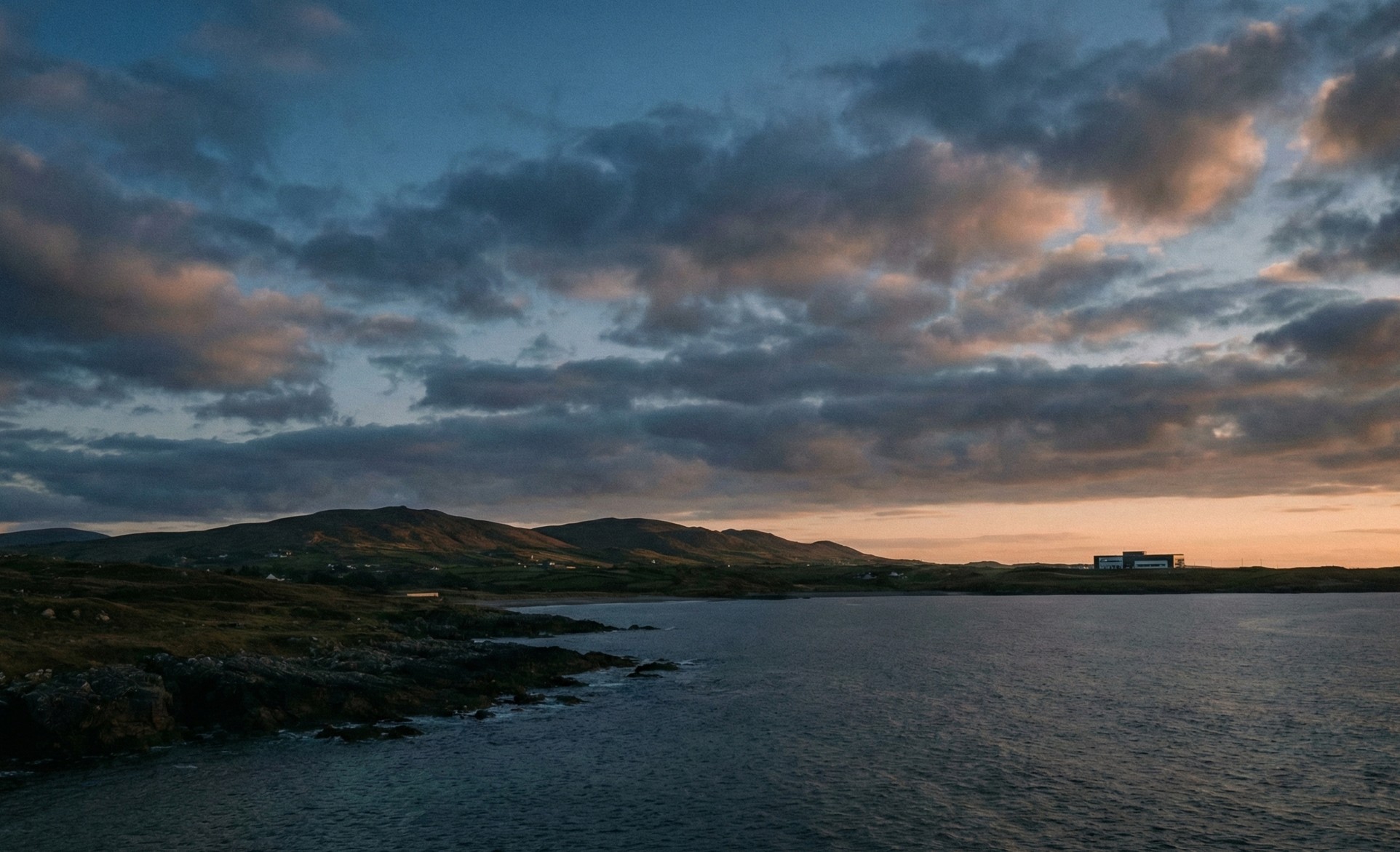Coastal landscape at sunset with rugged shoreline, calm water, hills in the background, and a modern building on the horizon.