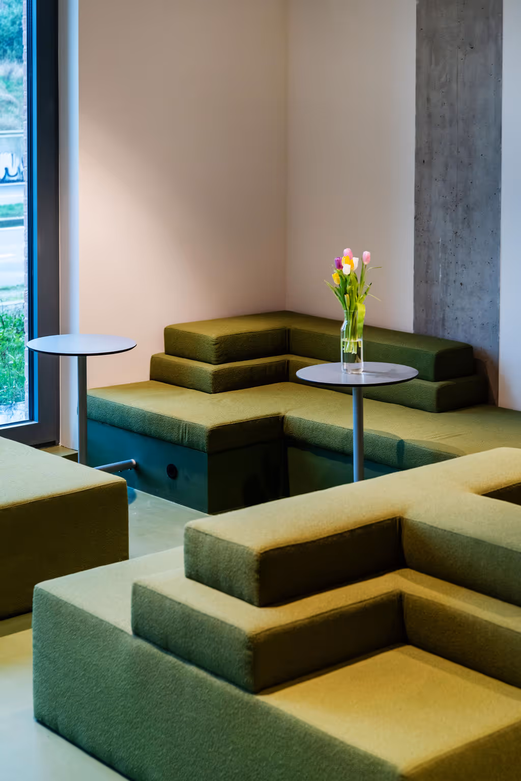 Modern indoor seating area with green tiered couches, round tables, and a vase of colorful tulips on one table.