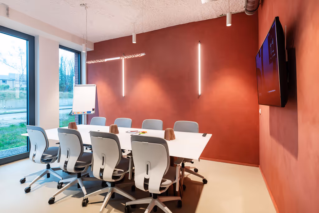 Modern conference room with white table, eight gray swivel chairs, a wall-mounted TV, two vertical LED light strips, and a flip chart near large windows.