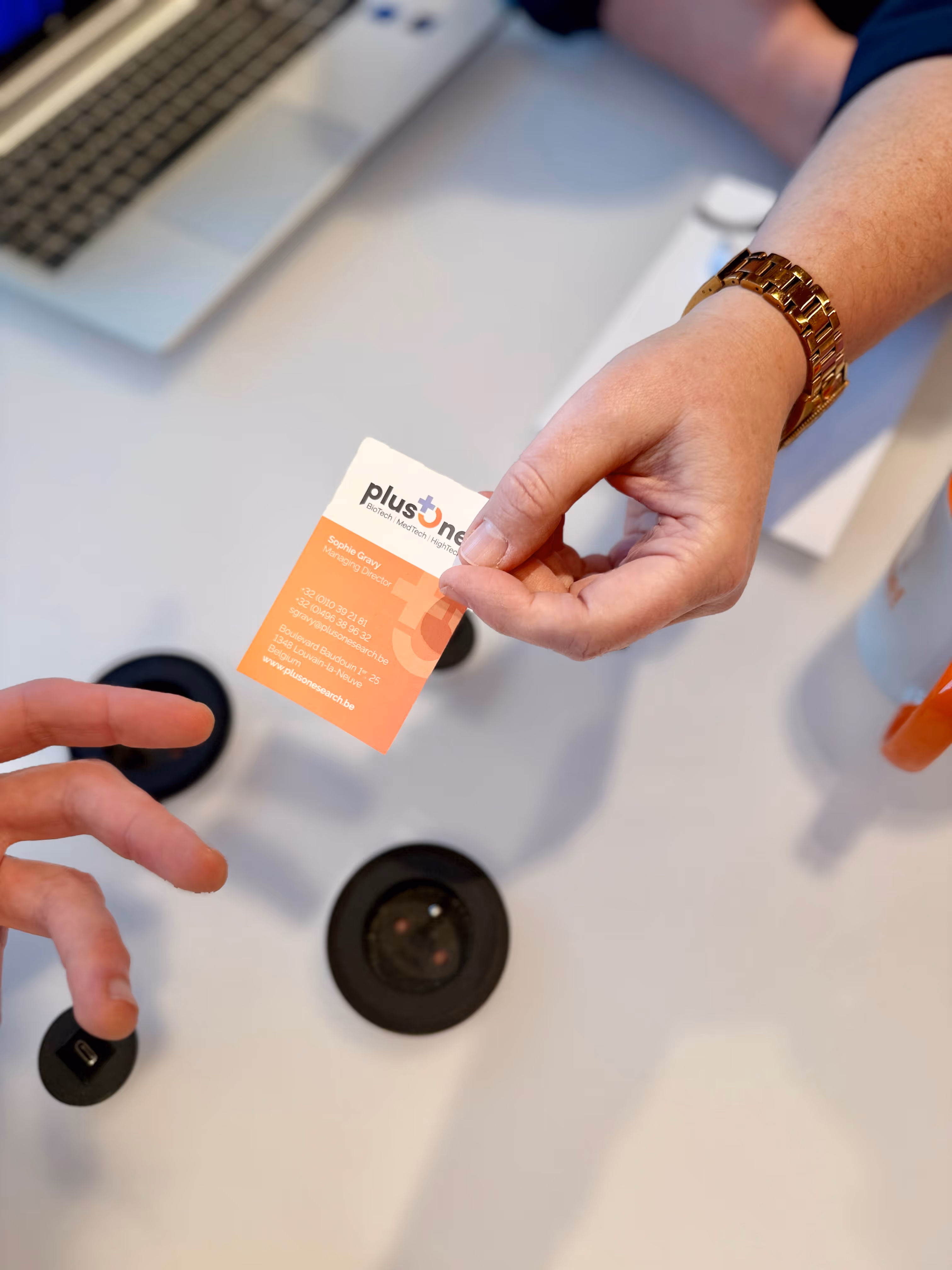 Person wearing a gold watch handing over a plusOne business card to another person across a white table with a laptop.