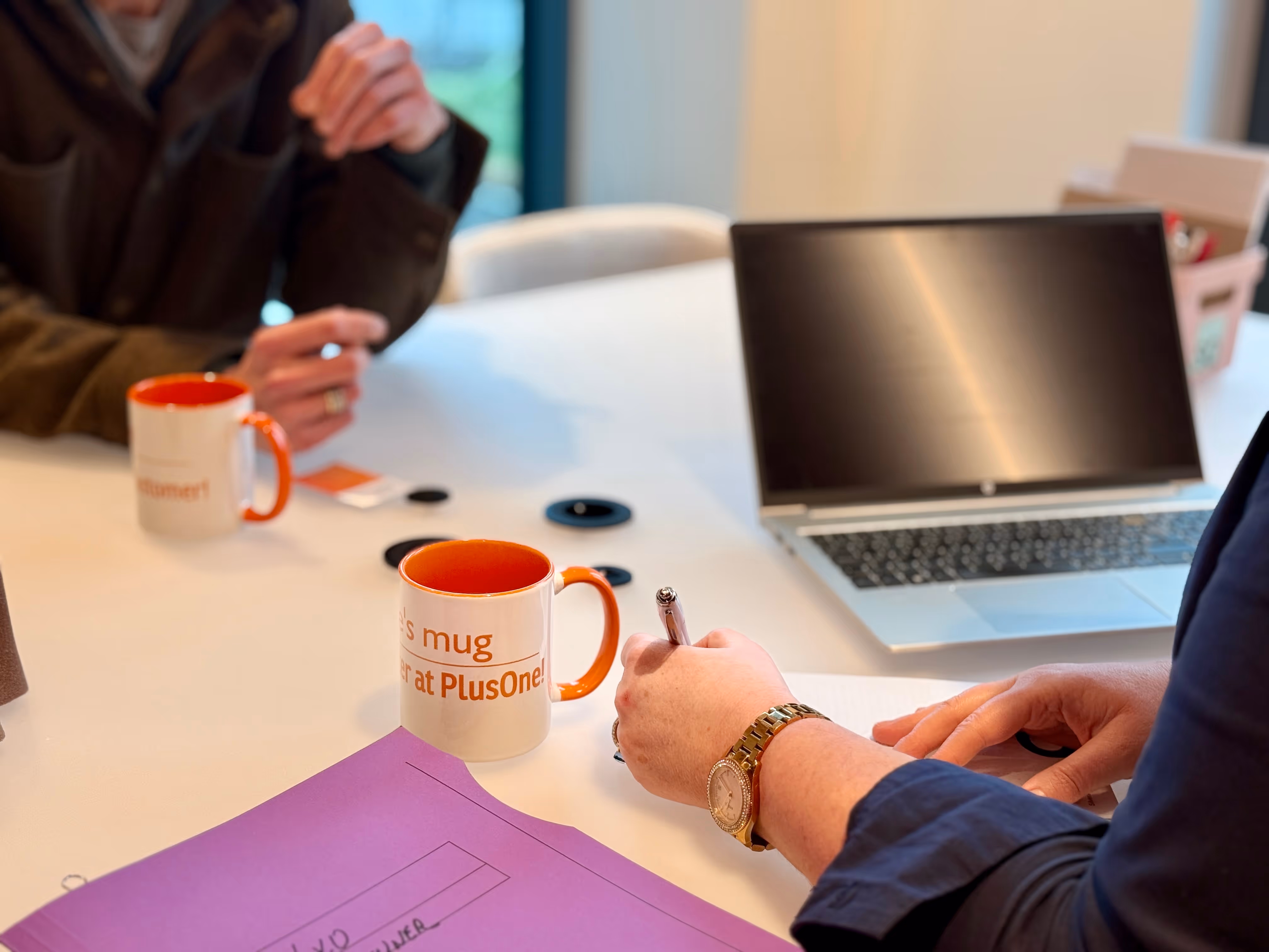 Office desk near window with a closed HP laptop, orange figurine with arms raised, eyeglasses, pens, and purple folders.