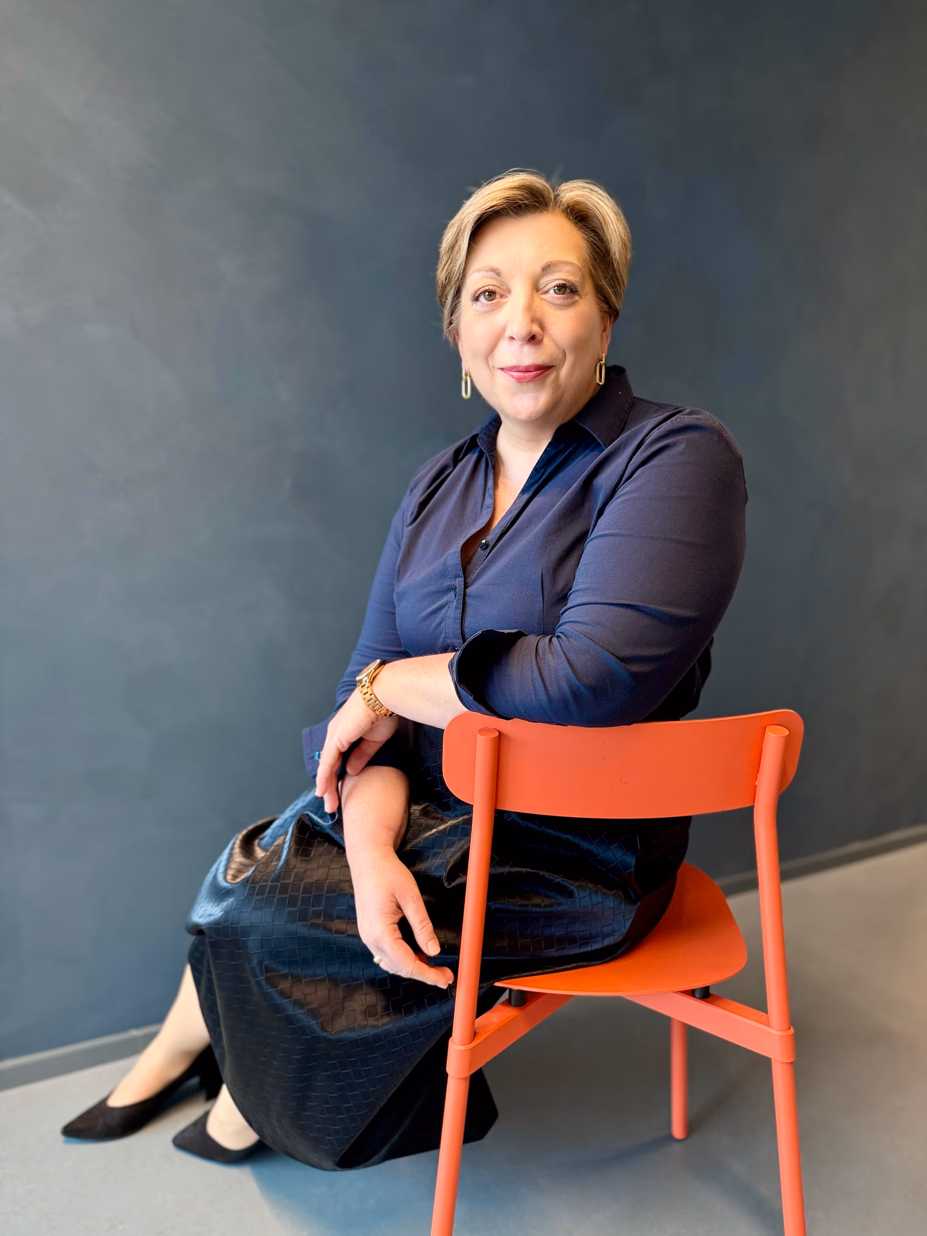 Middle-aged woman with short blonde hair wearing a blue shirt, seated on an orange chair against a blue background.
