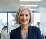 Femme d'affaires souriante aux cheveux gris courts, portant un blazer bleu et un collier, dans un bureau moderne avec des collègues en arrière-plan.
