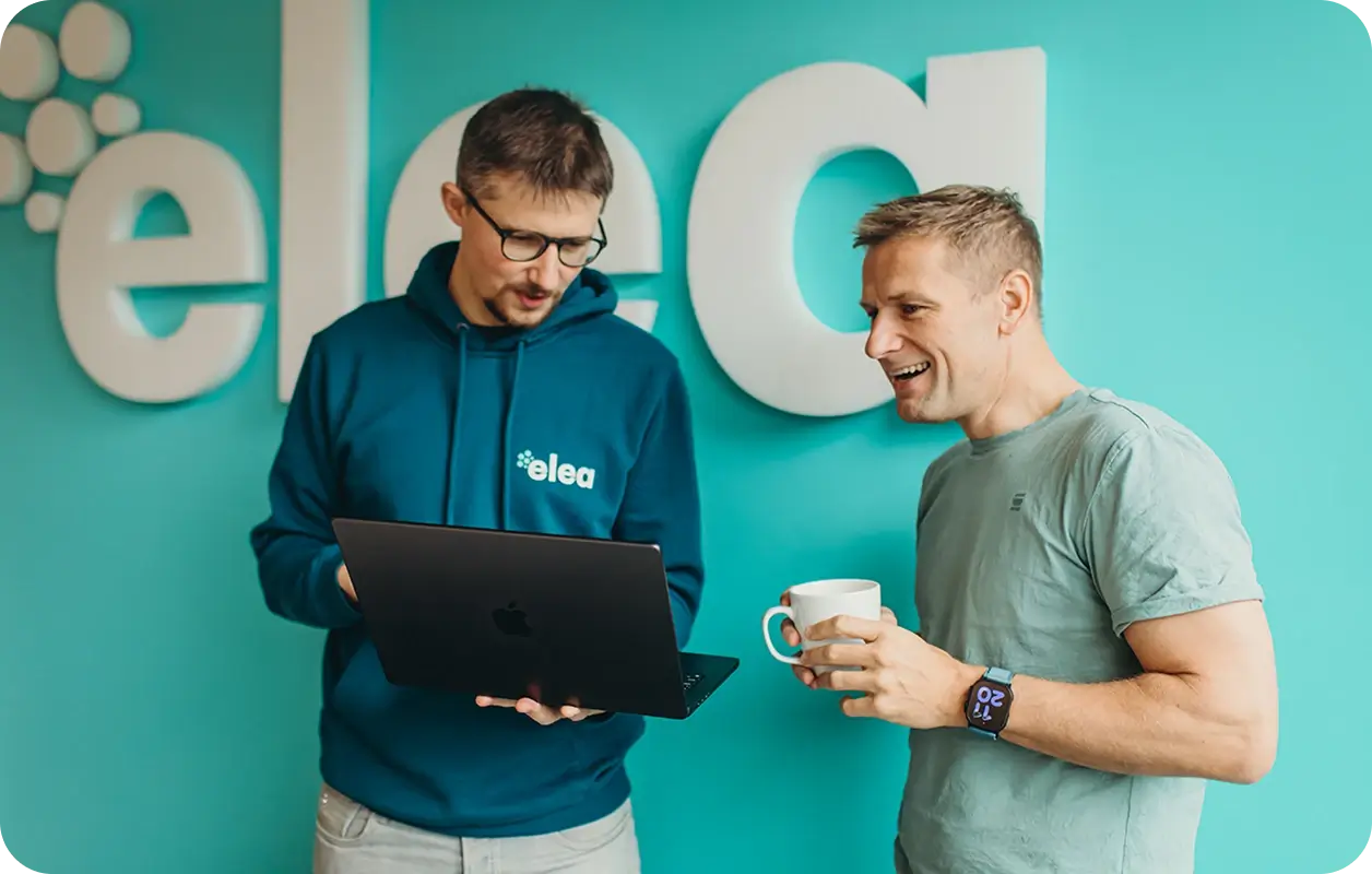 Two men standing in front of a turquoise wall with large white letters, one wearing a blue elea hoodie holding a laptop, and the other smiling with a coffee mug and smartwatch.