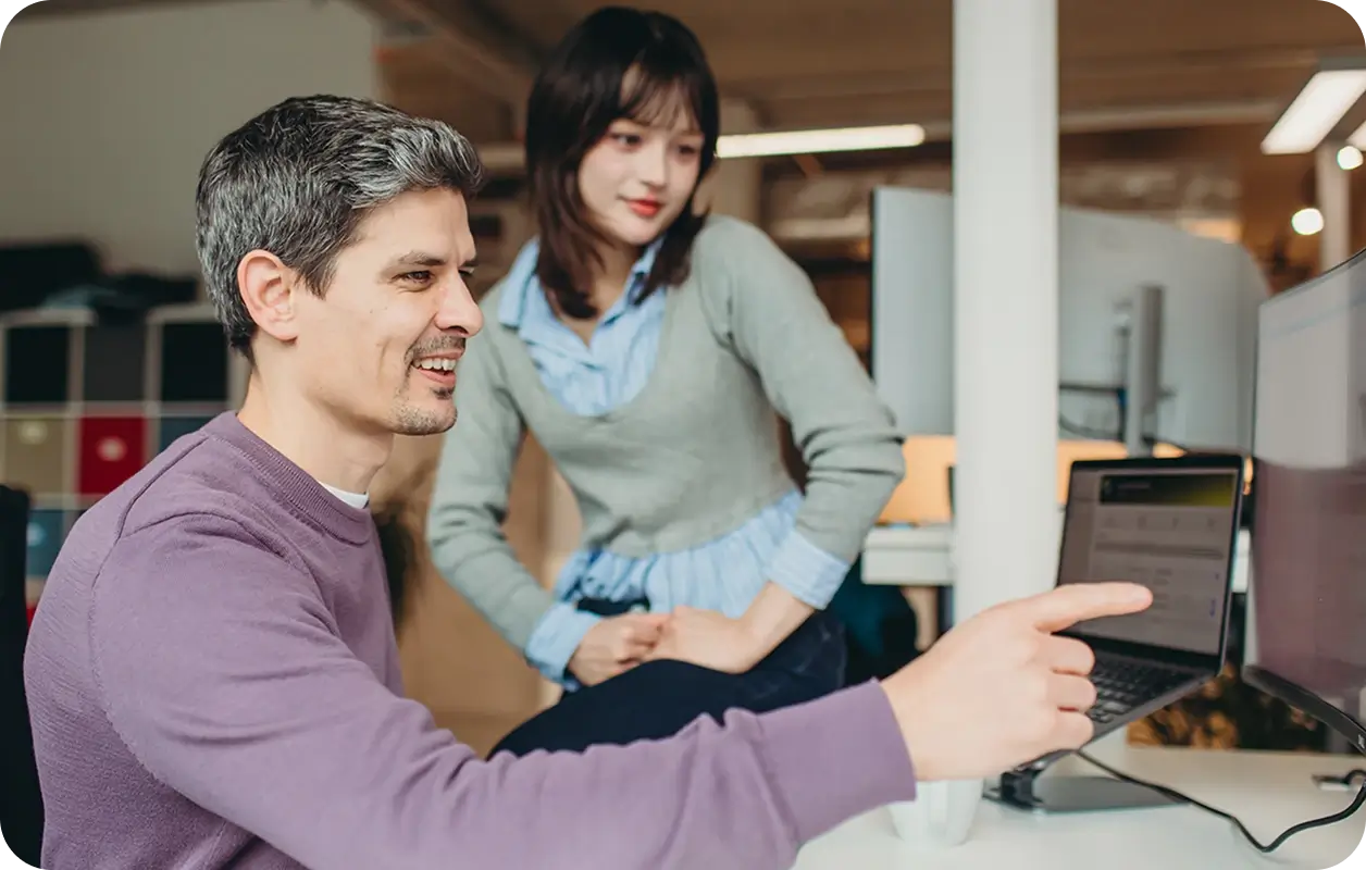 Man in a purple sweater pointing at a computer screen while a woman in a gray sweater and blue shirt watches attentively in a modern office.