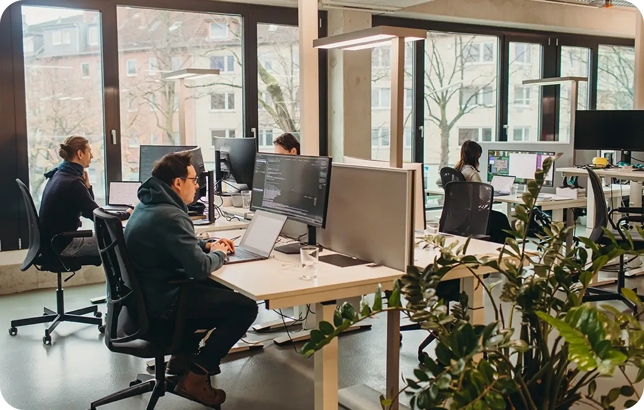 People working on laptops and desktop computers in a modern office with large windows and indoor plants.
