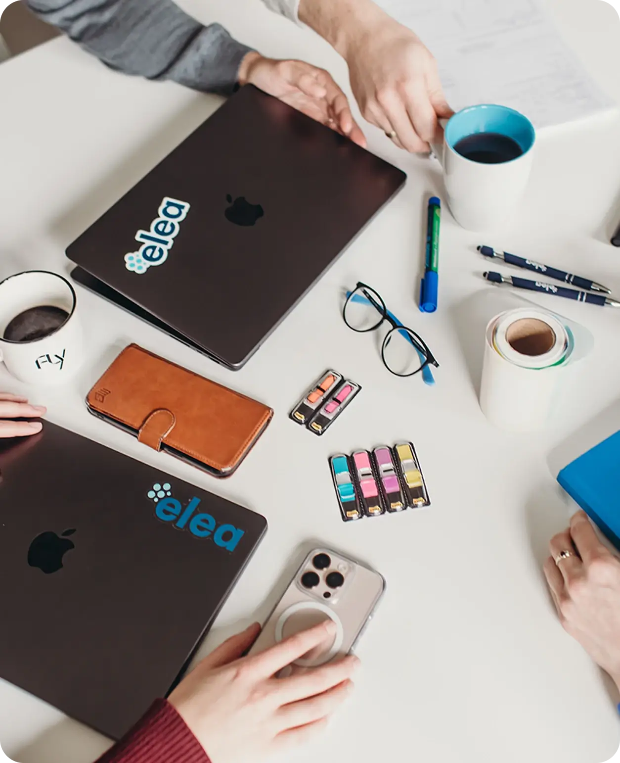 Arbeitsplatz mit zwei schwarzen Laptops, einem Smartphone, einer braunen Handyhülle, Brille, bunten Markierern, Stiften, Tassen und Händen von drei Personen, die sich über den Tisch beugen.